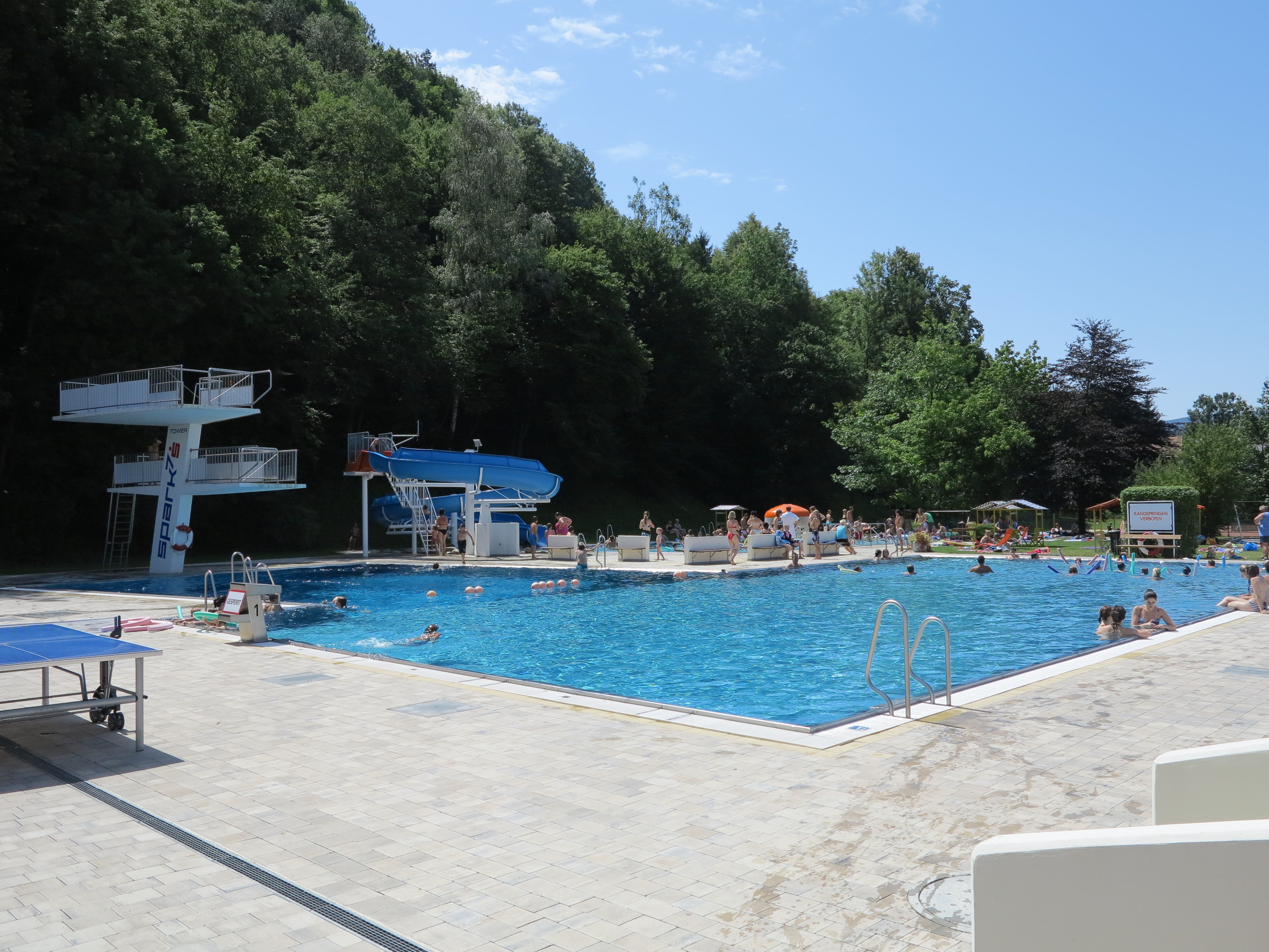 Venkovní bazén se skokanskou plošinou, skluzavkou a spoustou koupajících se za slunečného dne.