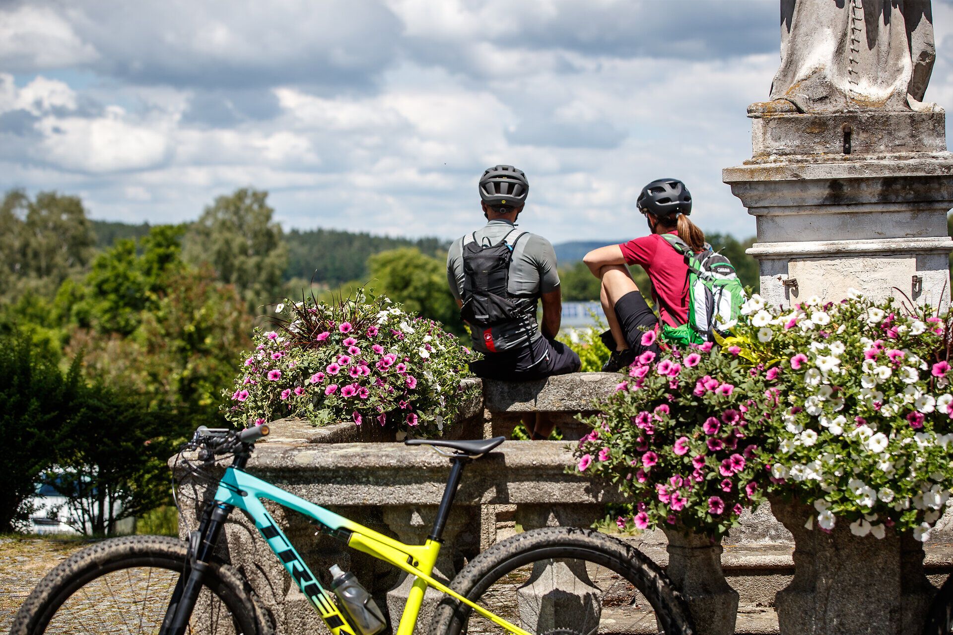 Dva cyklisté na horských kolech si užívají zaslouženého odpočinku uprostřed rozkvetlé krajiny. Toto místo obklopené barevnými květinami a čerstvým horským vzduchem nabízí dokonalou kulisu pro dobrodružství v přírodě.