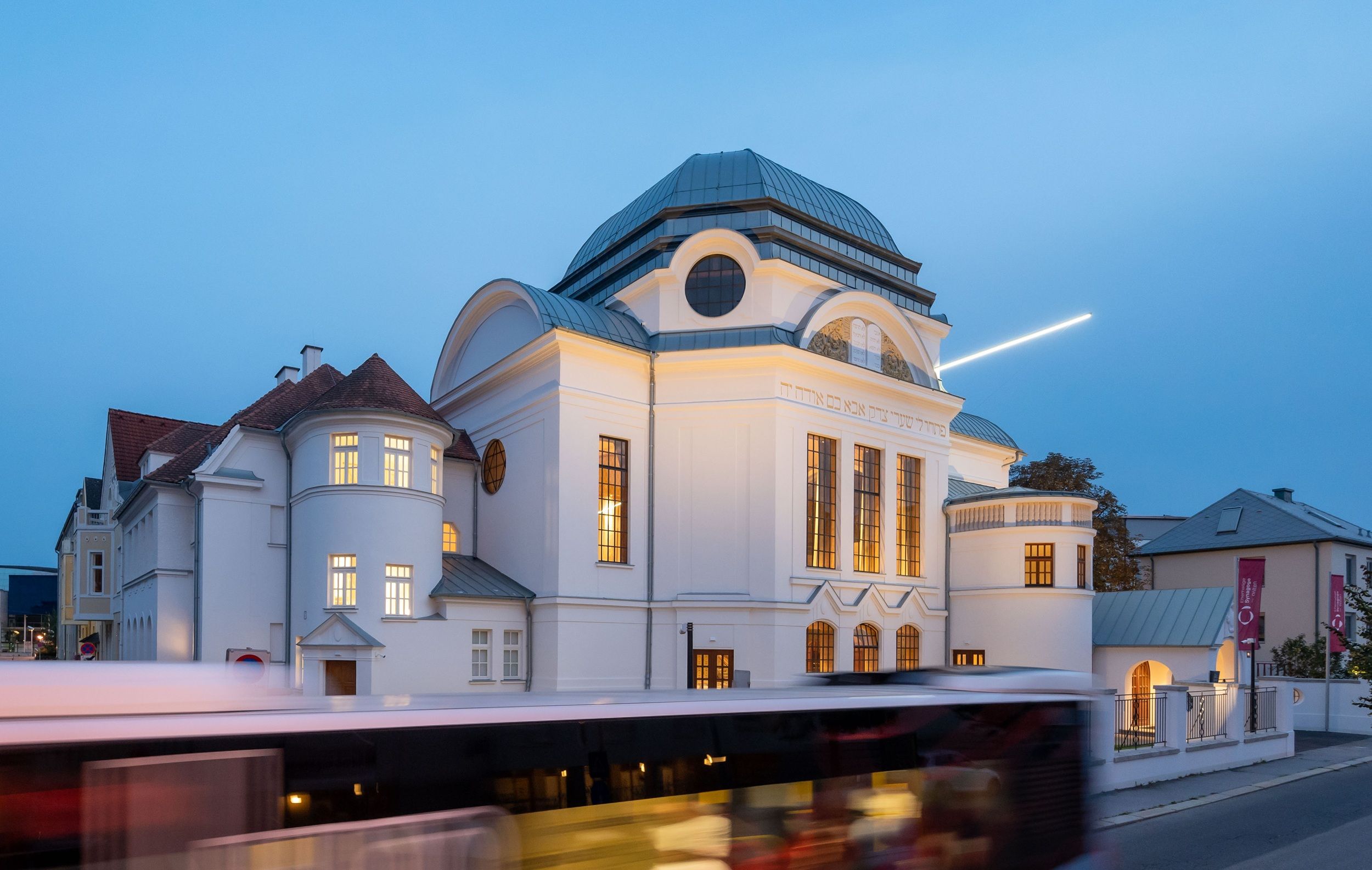 Bývalá synagoga v St. Pöltenu za soumraku s osvětlenými okny a projíždějícím autobusem.