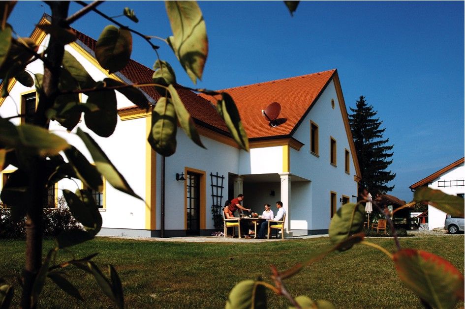 Bílý dům s červenou střechou, obklopený zeleným trávníkem a stromy. Venku sedí lidé u stolu.