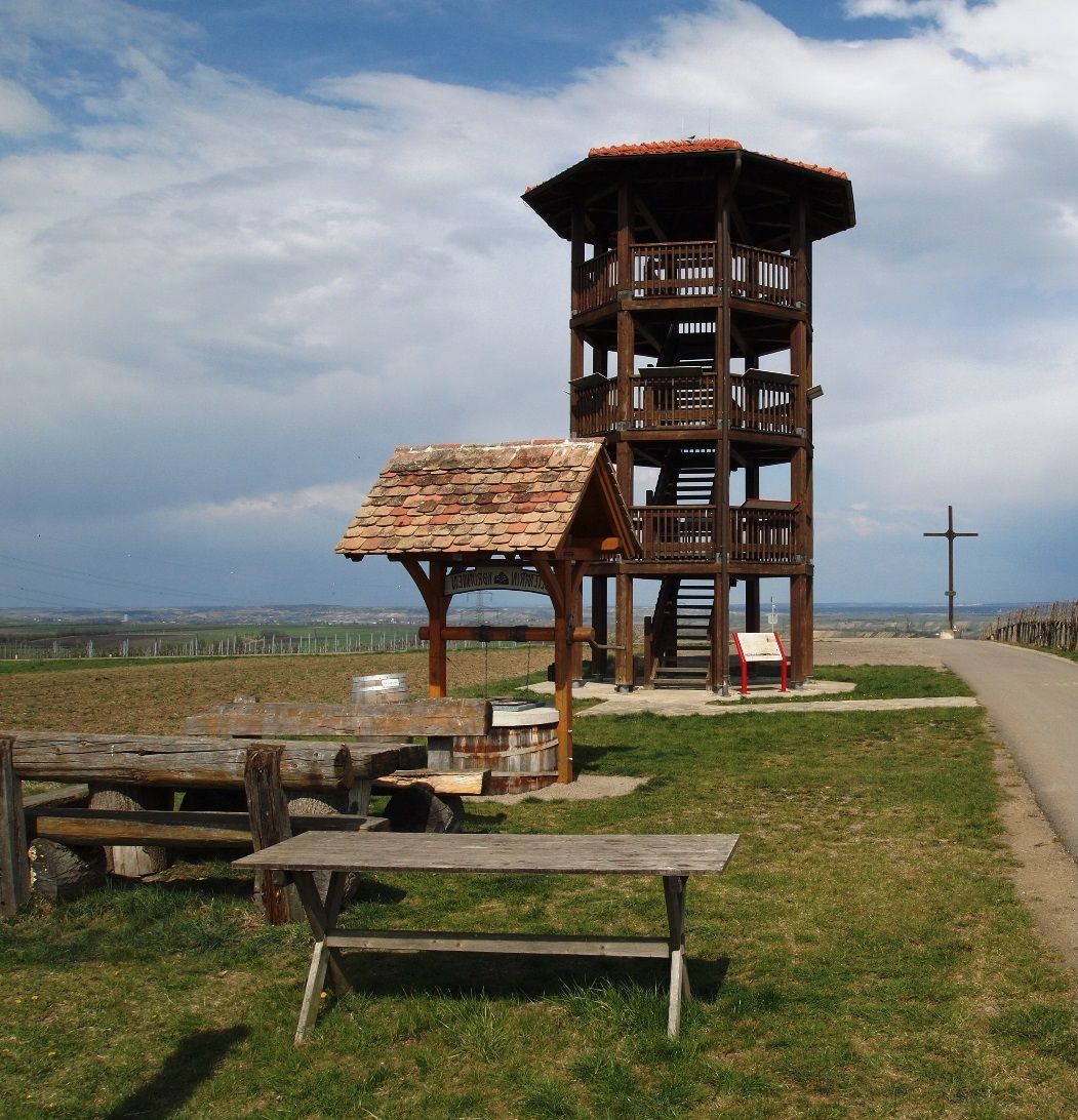 Dřevěná rozhledna na louce s lavičkami a křížem v pozadí.