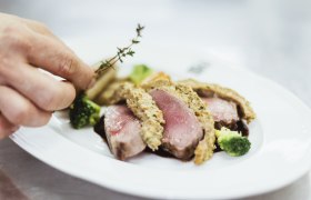 Detailní záběr na růžový pokrm z pečeného masa s bylinkovou krustou, brokolicí a rukou přidávající tymián.