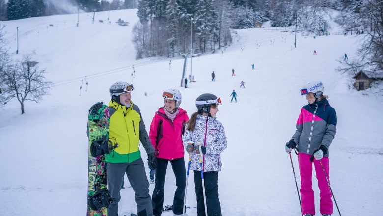 Skupina lyžařů a snowboardistů na zasněžen&eacute; sjezdovce s lesem v pozad&iacute;.