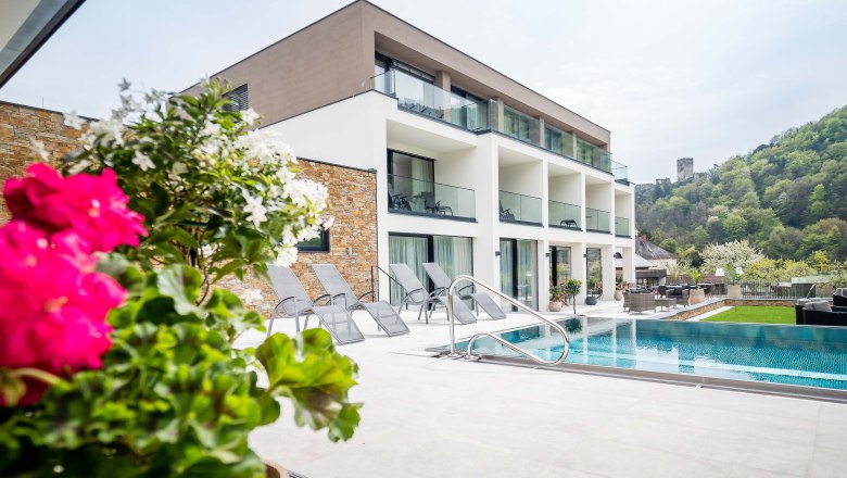 Moderní budova s bazénem a lehátky, obklopená zelenou krajinou a hradem v pozadí.