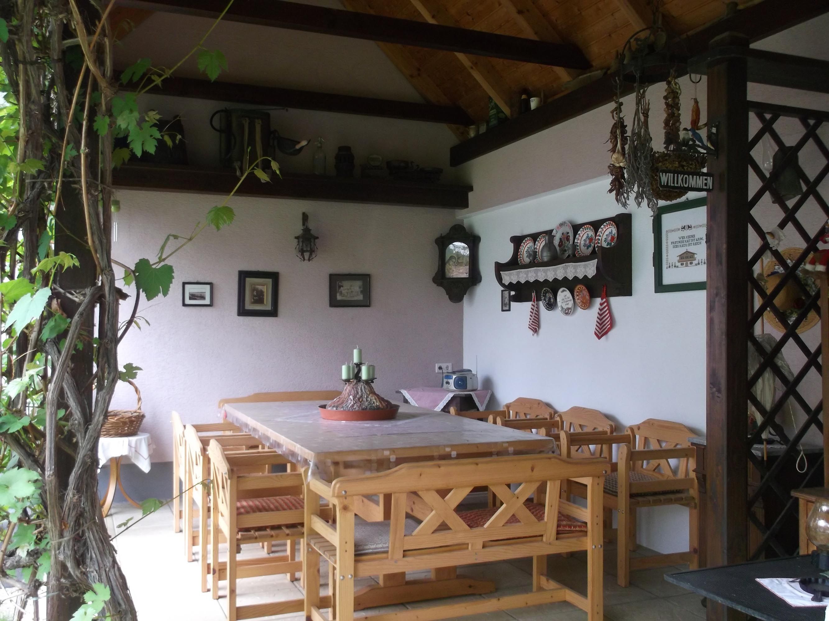 Zastřešená terasa se stolem a židlemi, v pozadí zeď s různými nástěnnými dekoracemi.