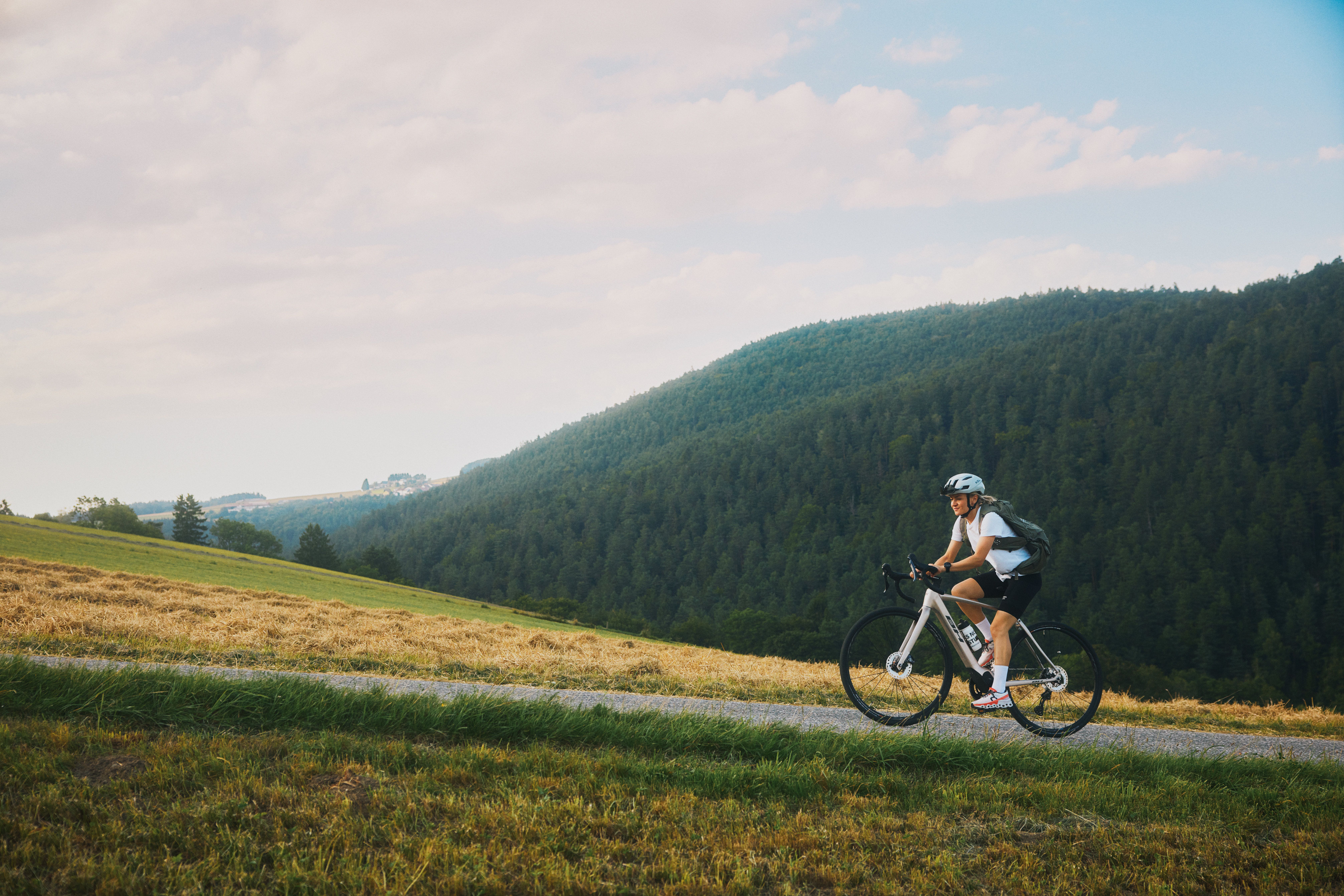 Cyklista jede po úzké stoupající stezce se zalesněnými kopci v pozadí.
