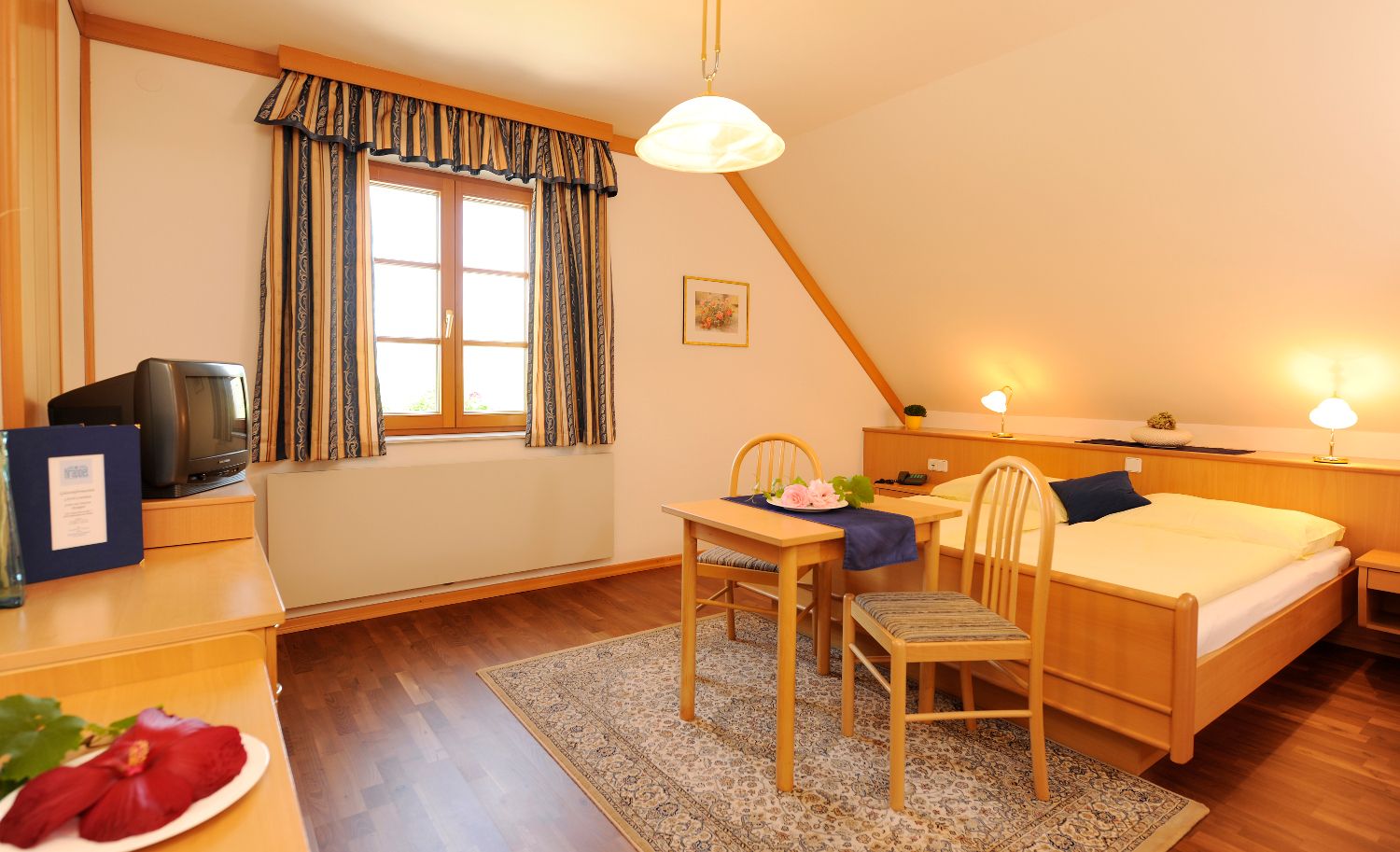 Útulný pokoj pro hosty s dřevěným nábytkem, manželskou postelí, stolkem a židlemi, TV a oknem se závěsy.
