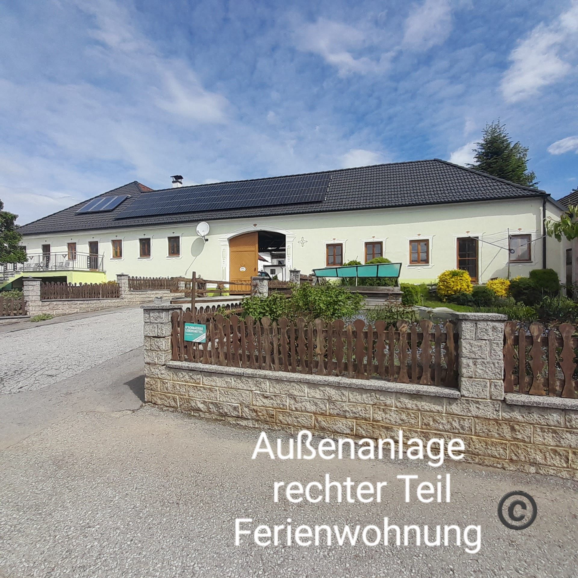 Velká budova se žlutou fasádou a černou střechou, obklopená zahradou a dřevěným plotem. Obloha je modrá s několika mraky.