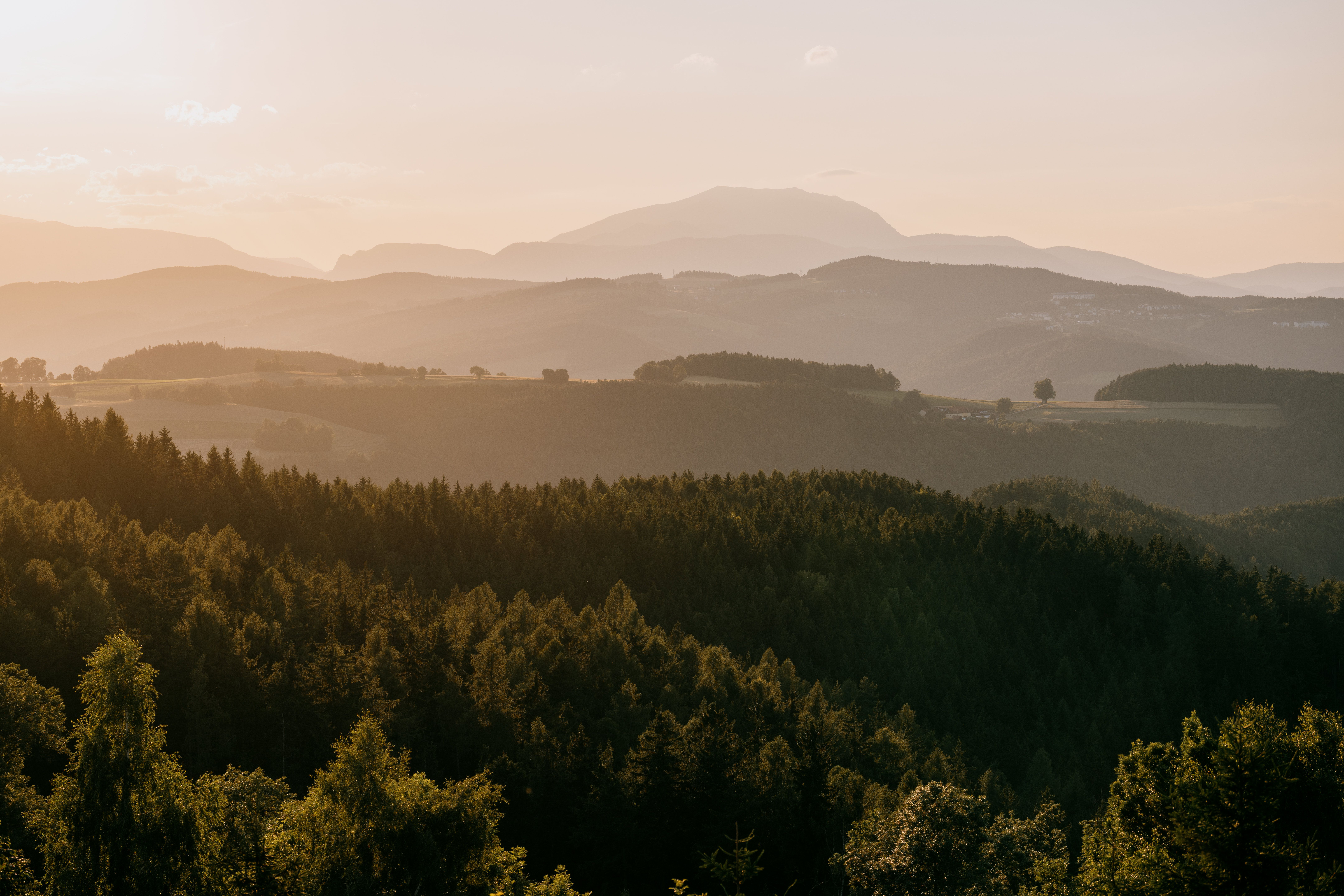 Západ slunce nad zalesněnou kopcovitou krajinou se zasněženou horou v pozadí.