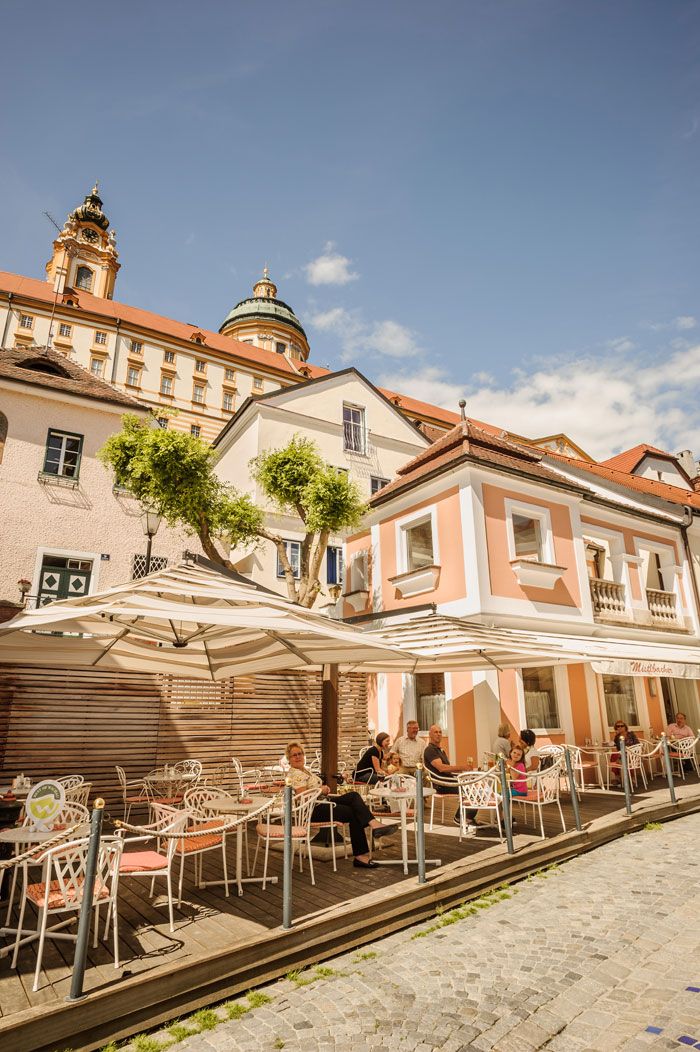 Venkovní prostor kavárny se slunečníky a hosty, v pozadí historická budova s kopulí.