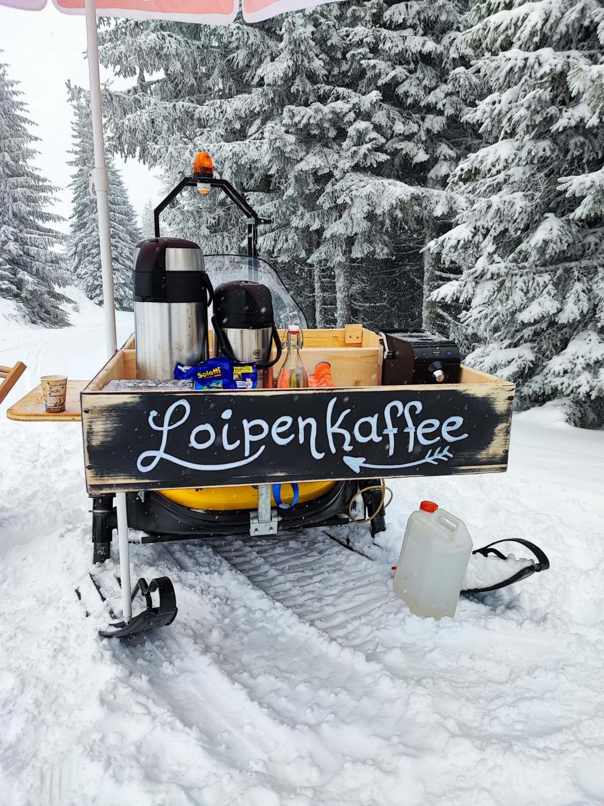 Mobilní stánek s kávou na sněhu s nápisem "Loipenkaffee".