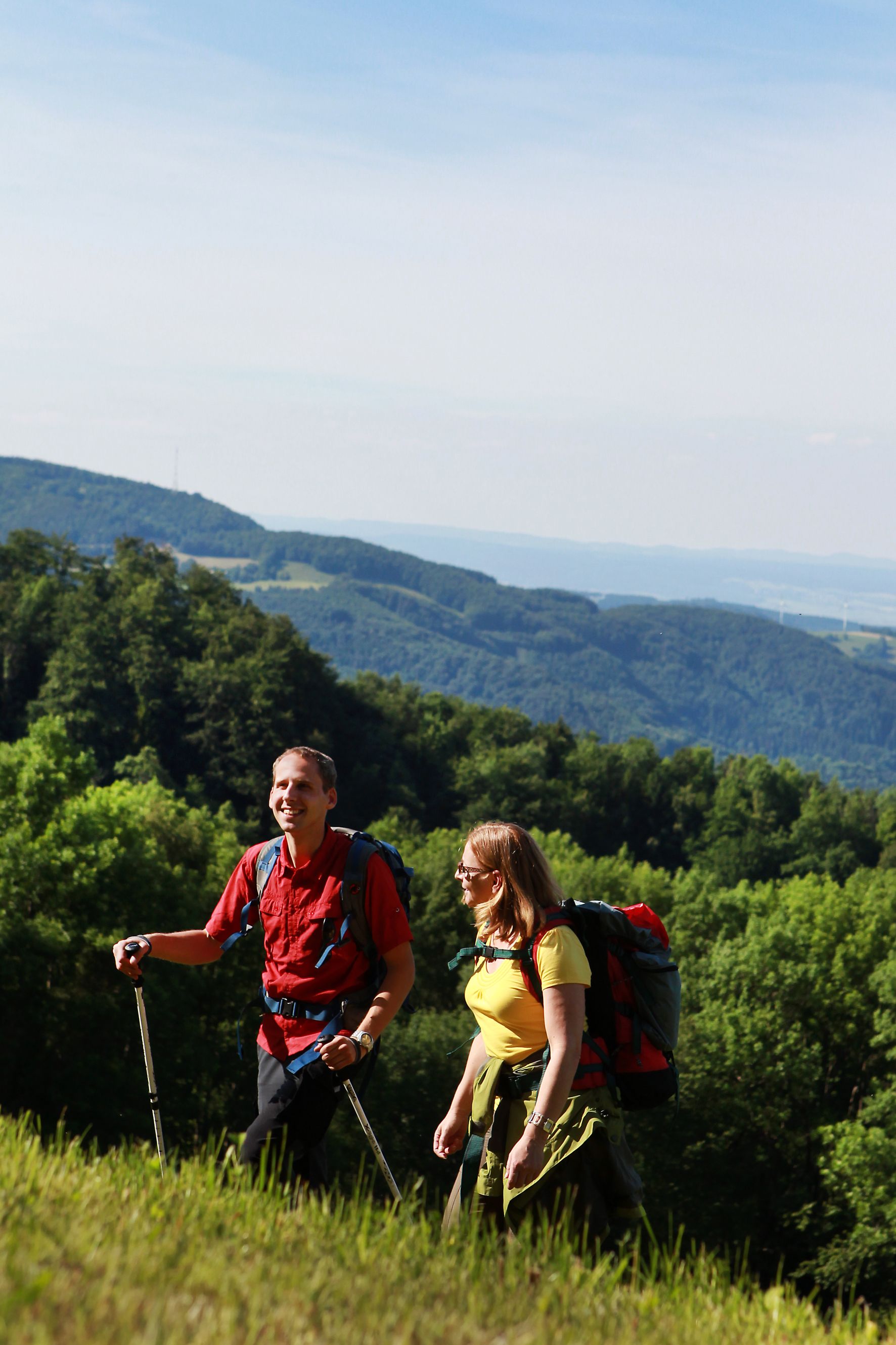 Dva turisté s batohy a hůlkami na zeleném kopci se zalesněnými horami v pozadí.