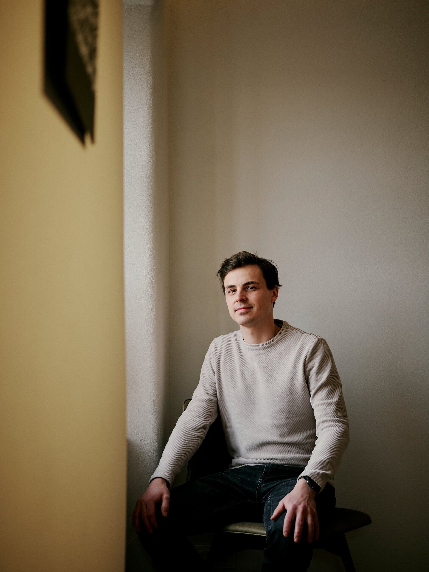 Osoba sedí na stoličce v prostém interiéru a dívá se do kamery.