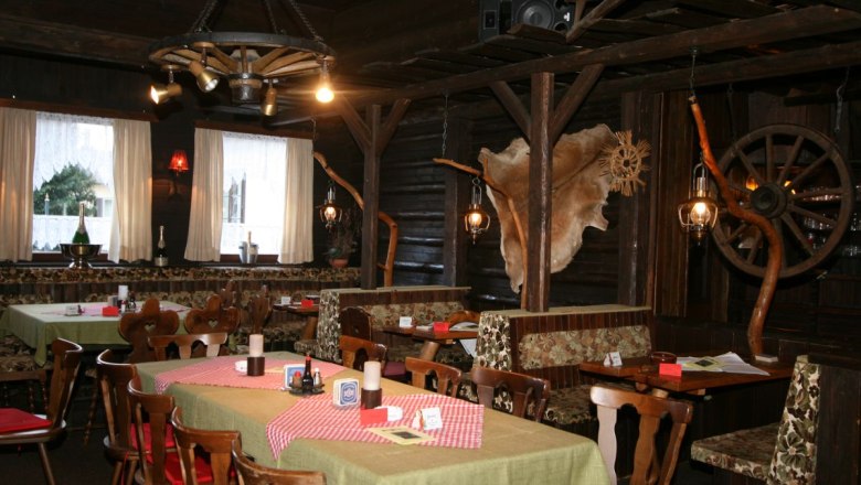 Rustik&aacute;ln&iacute; restaurace s dřevěn&yacute;m n&aacute;bytkem, kostkovan&yacute;mi ubrusy a dekorativn&iacute;mi prvky, jako jsou kola vozů a zv&iacute;řec&iacute; kože&scaron;iny na stěn&aacute;ch.