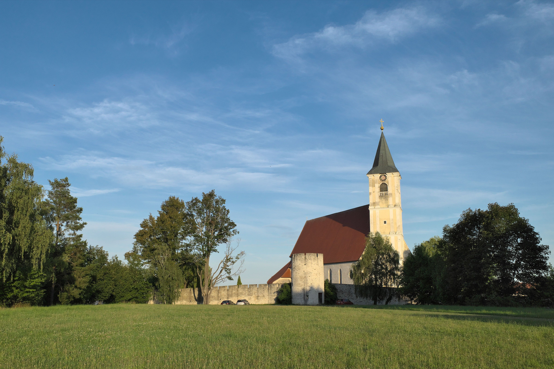 Kostel s věží a červenou střechou na louce, obklopený stromy a zdí.