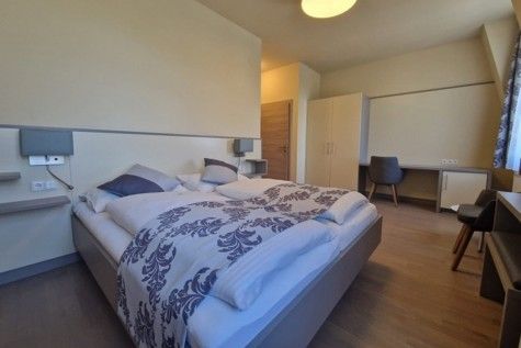Moderně zařízený hotelový pokoj s manželskou postelí, psacím stolem a šatní skříní.