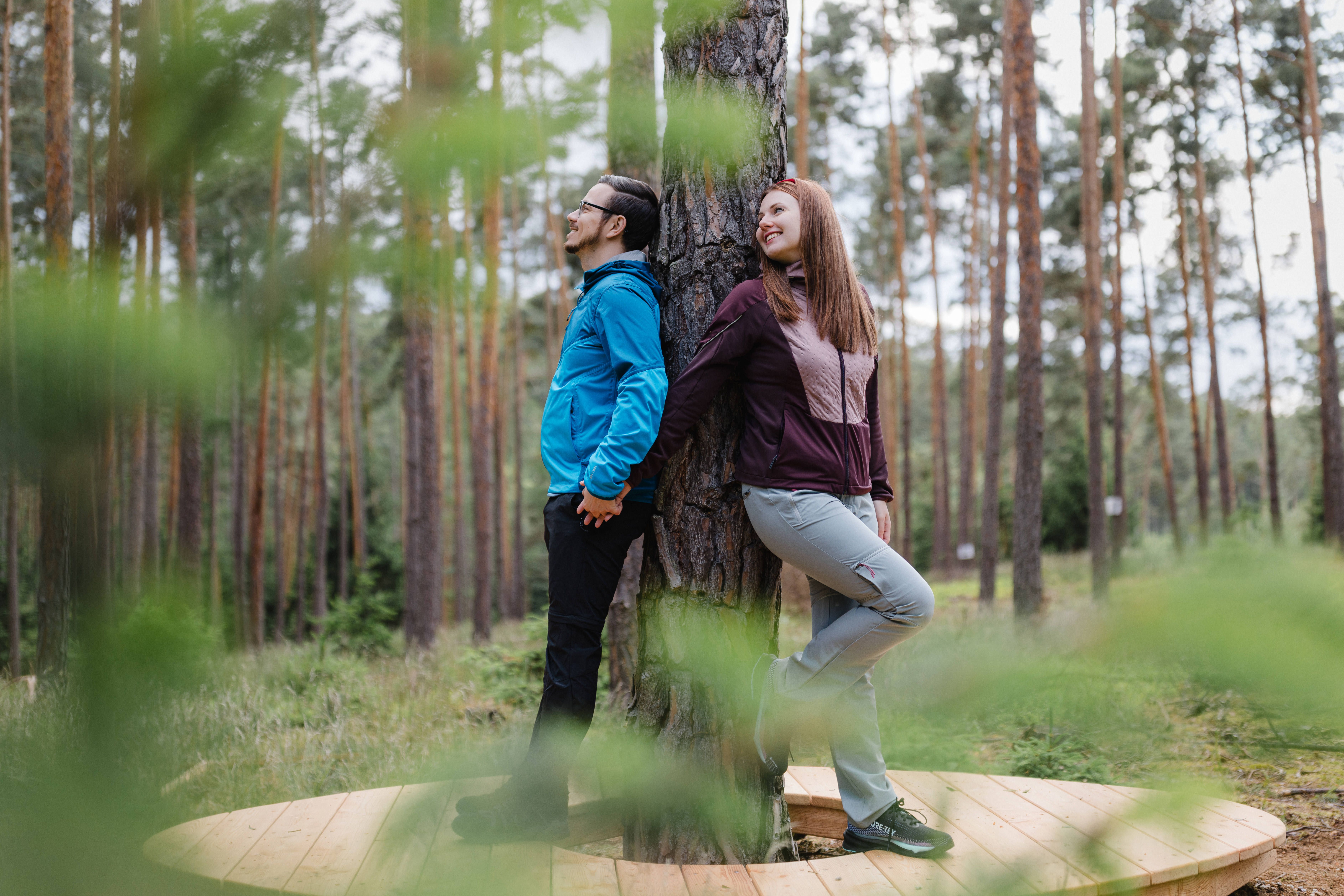 Dva lidé opření o strom v lese, stojící na dřevěné plošině.