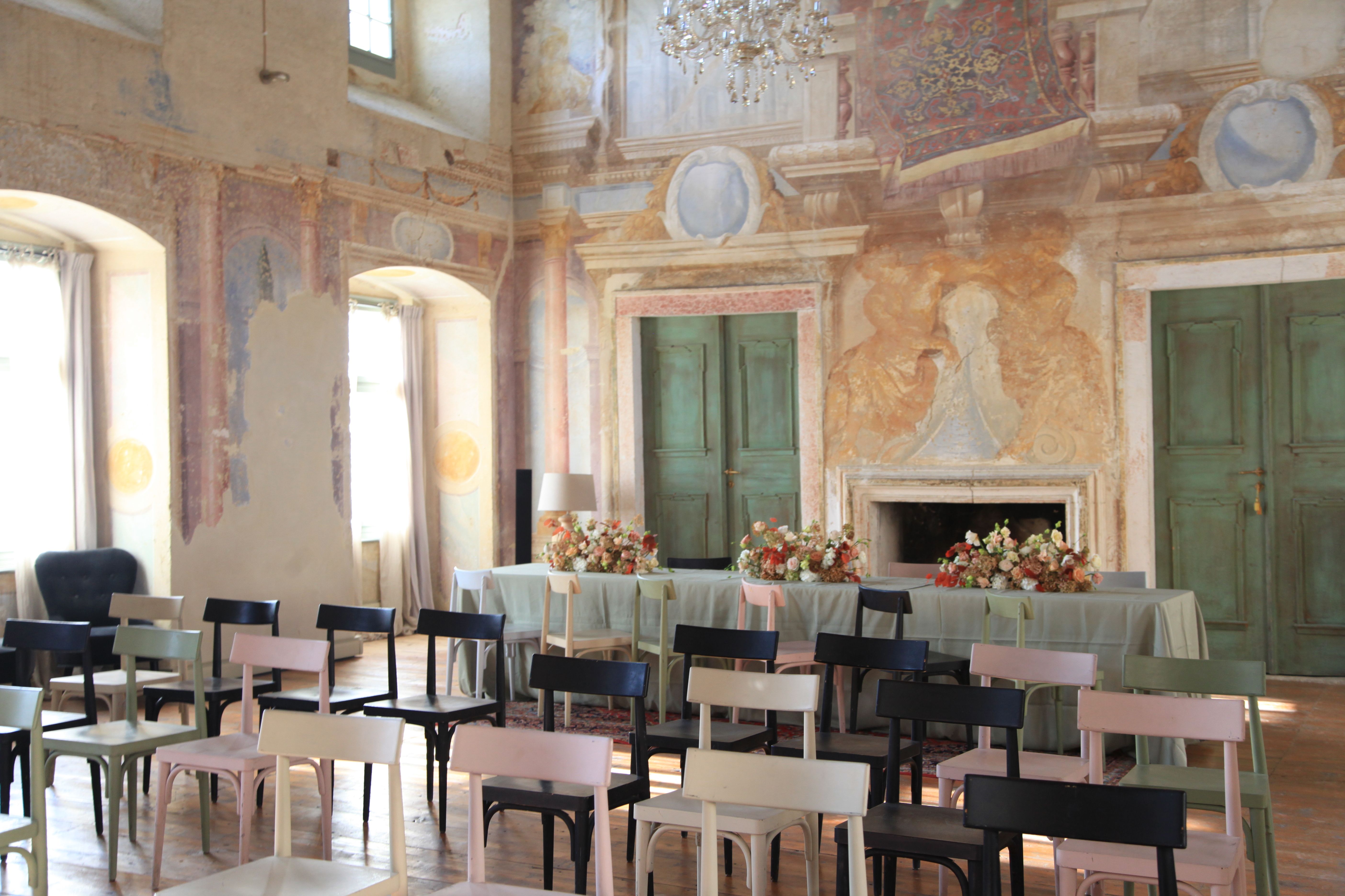 Slavnostně vyzdobený sál s malovanými stěnami, židlemi a stolem s květinami.