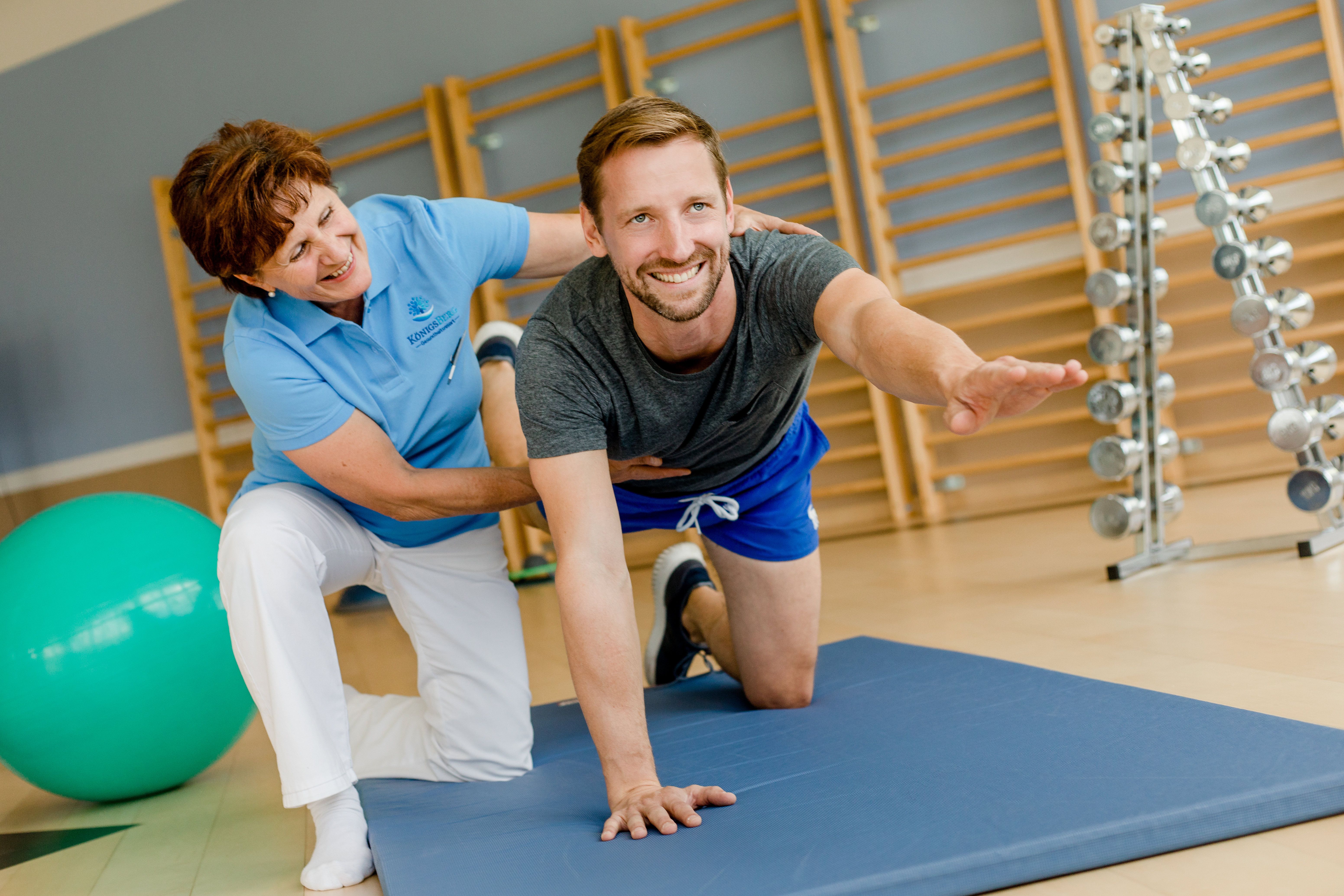Muž cvičí na podložce za podpory terapeuta ve fitness centru.