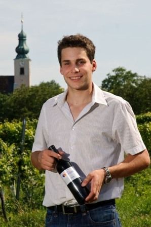 Muž stojí na vinici a drží láhev vína. V pozadí je vidět kostelní věž.