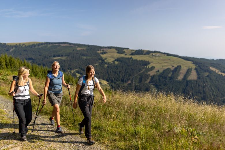 Tři ženy s turistickými holemi a batohy kráčejí po horské stezce obklopené zelenými kopci a lesy.