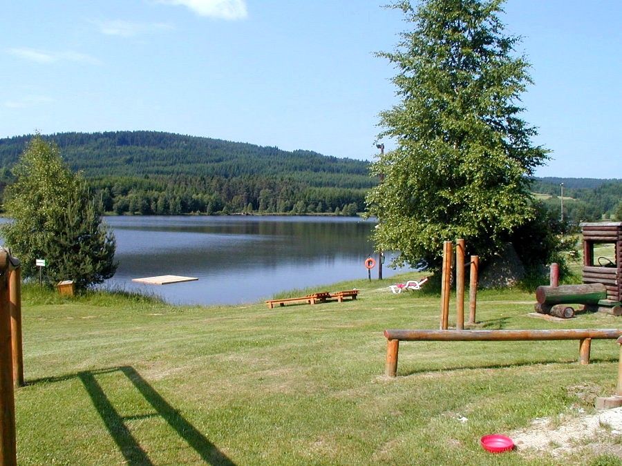 Klidné jezero s molem, obklopené zelenými kopci a stromy, s dětským hřištěm v popředí.