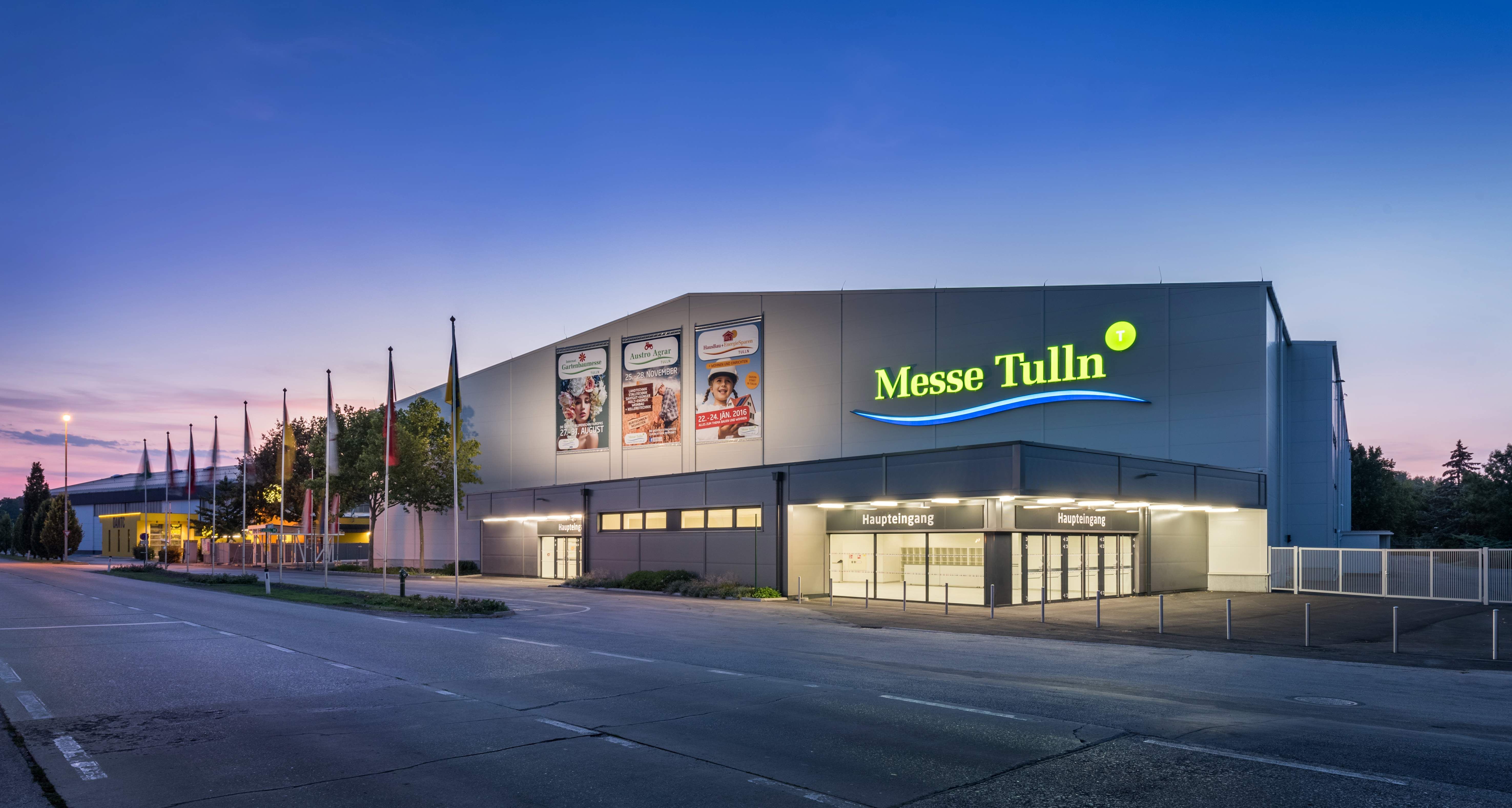 Vnější pohled na výstaviště Tulln za soumraku s osvětleným vchodem a vlajkami.