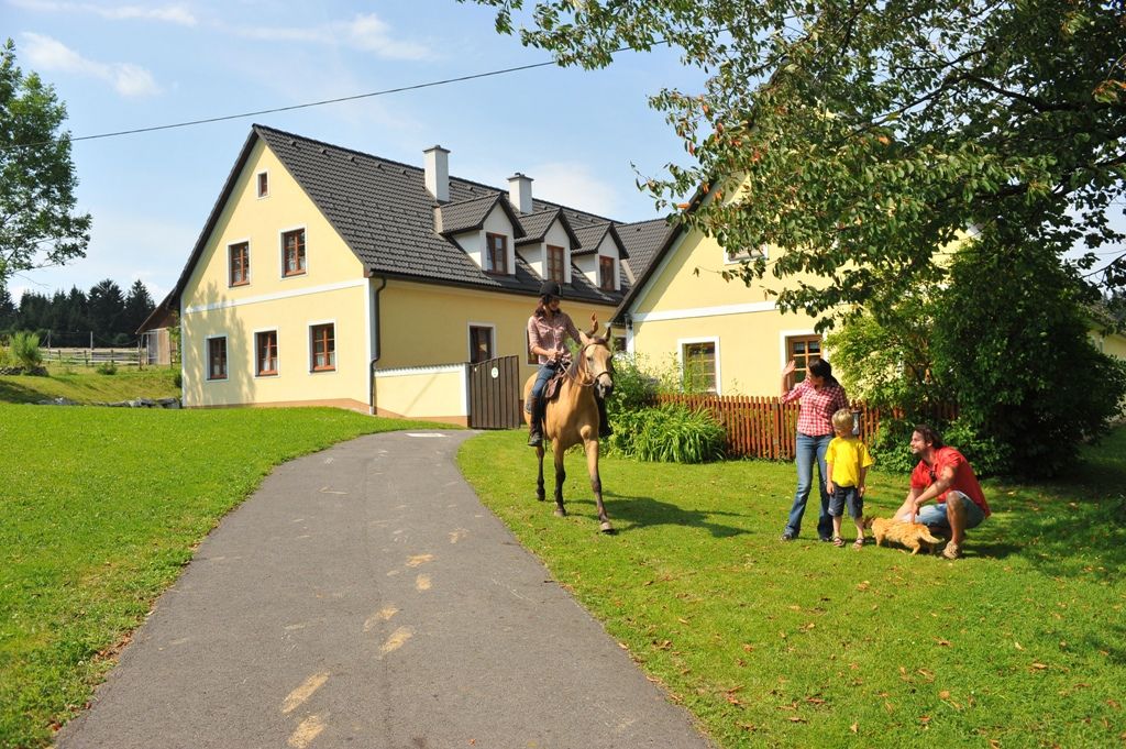 Jezdec na koni se blíží k rodině před žlutým domem na farmě.