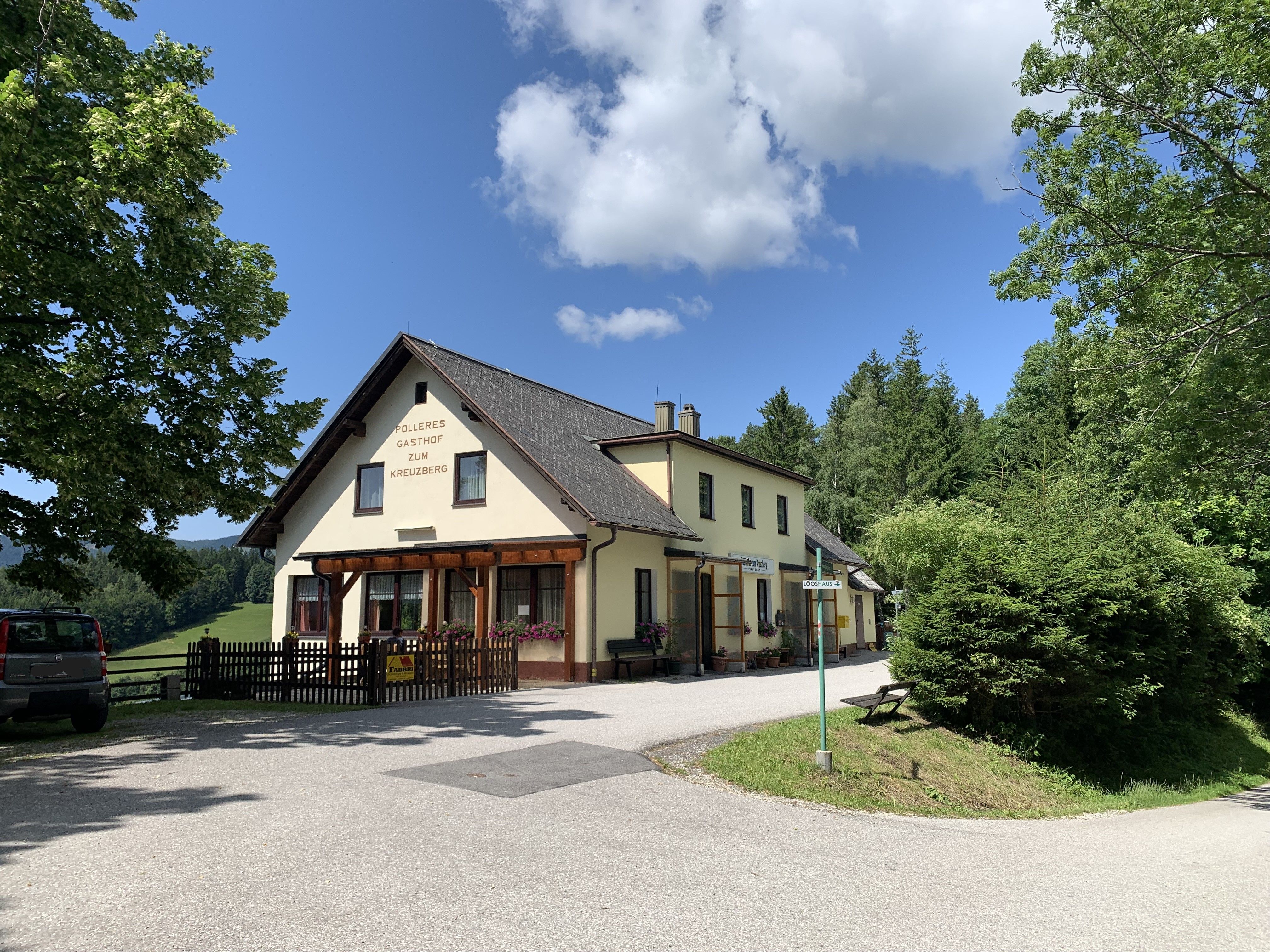 Žlutý hostinec s nápisem "Polleres Gasthof zum Kreuzberg" obklopený stromy a modrou oblohou.