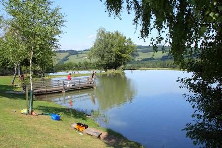 Klidné jezero s molem a rybáři, obklopené zelenými stromy a kopci v pozadí.
