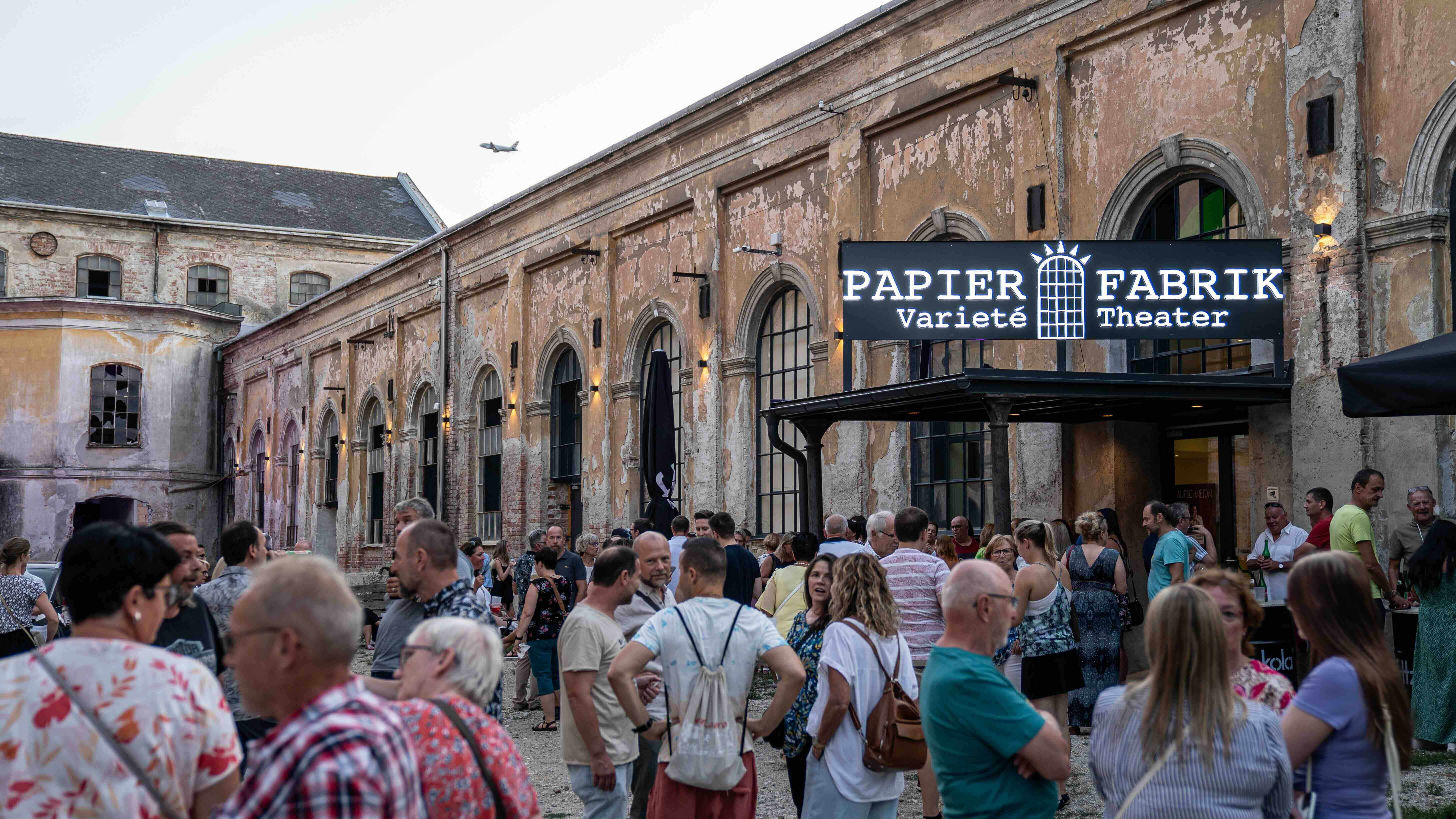 Dav lidí před starou továrnou na papír s nápisem "Papier Fabrik Varieté Theater".