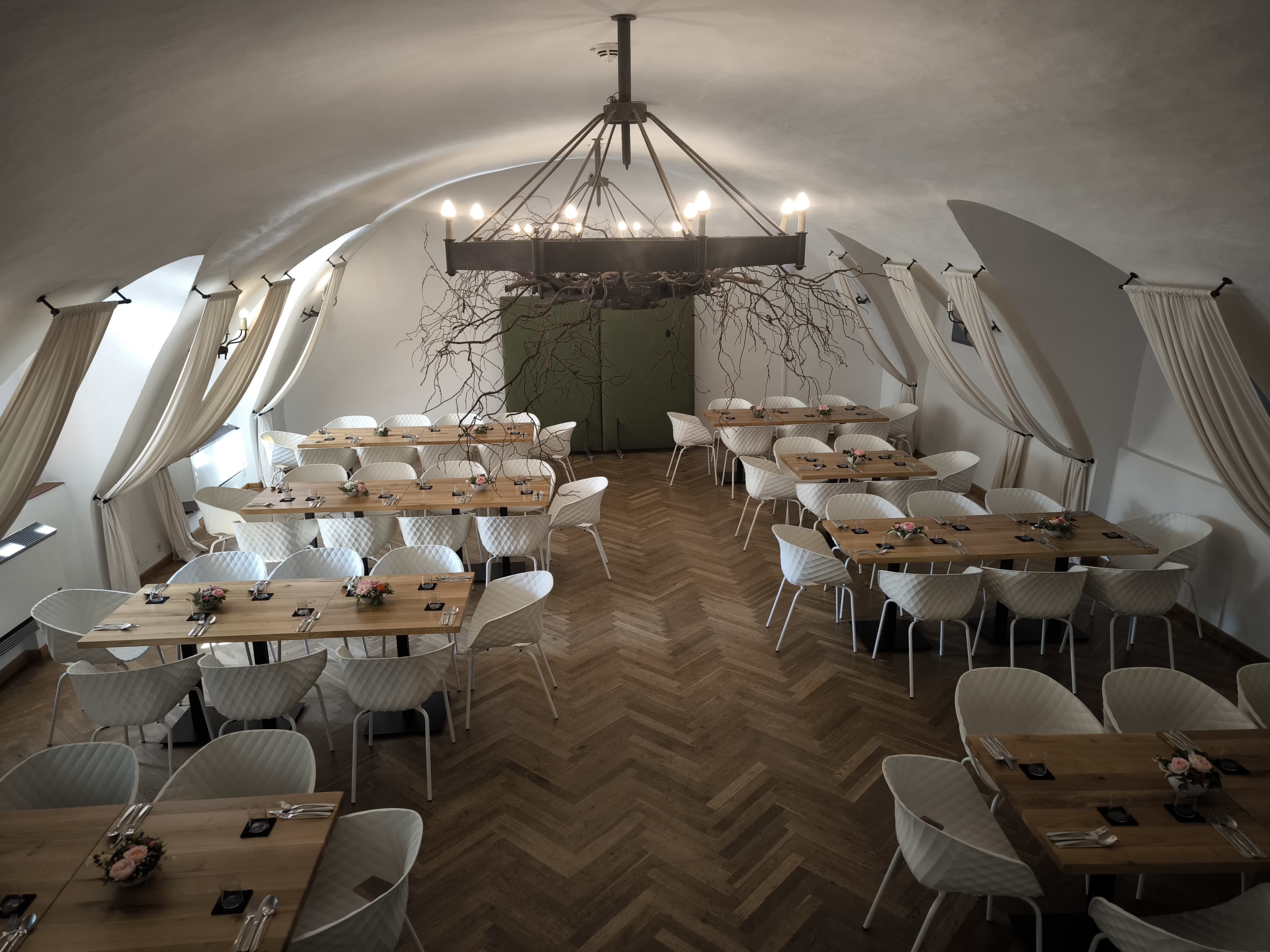 Elegantní sál s dřevěnými stoly, parketovou podlahou, bílými židlemi a železným lustrem.