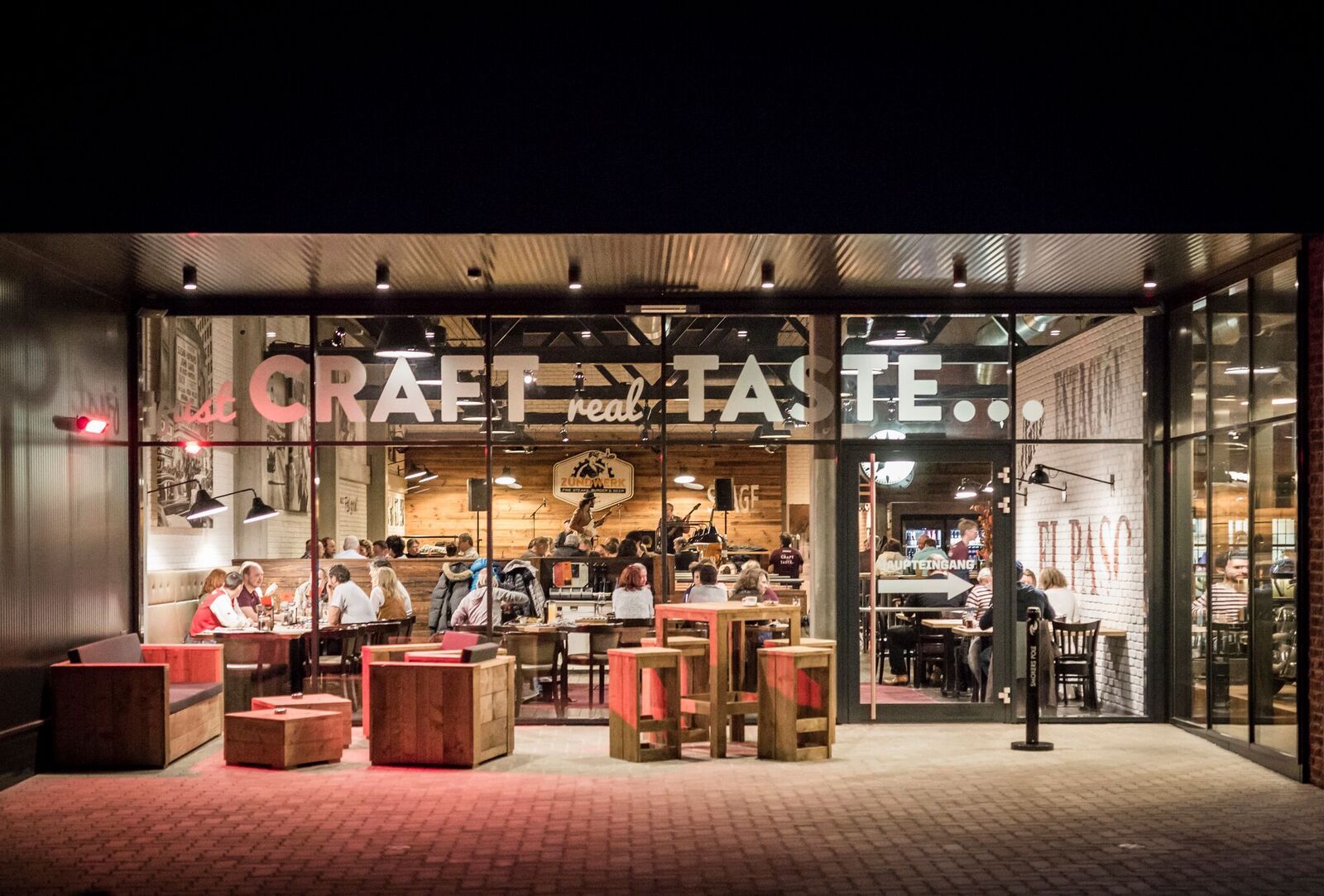 Pohled zvenčí na restauraci s proskleným průčelím, uvnitř sedí lidé u stolů.