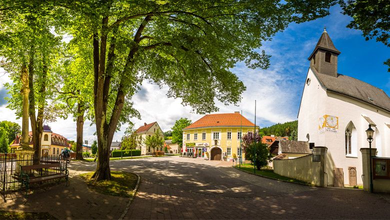 Panoramatický pohled na náměstí Feistritz s kostelem, stromy a historickými budovami.