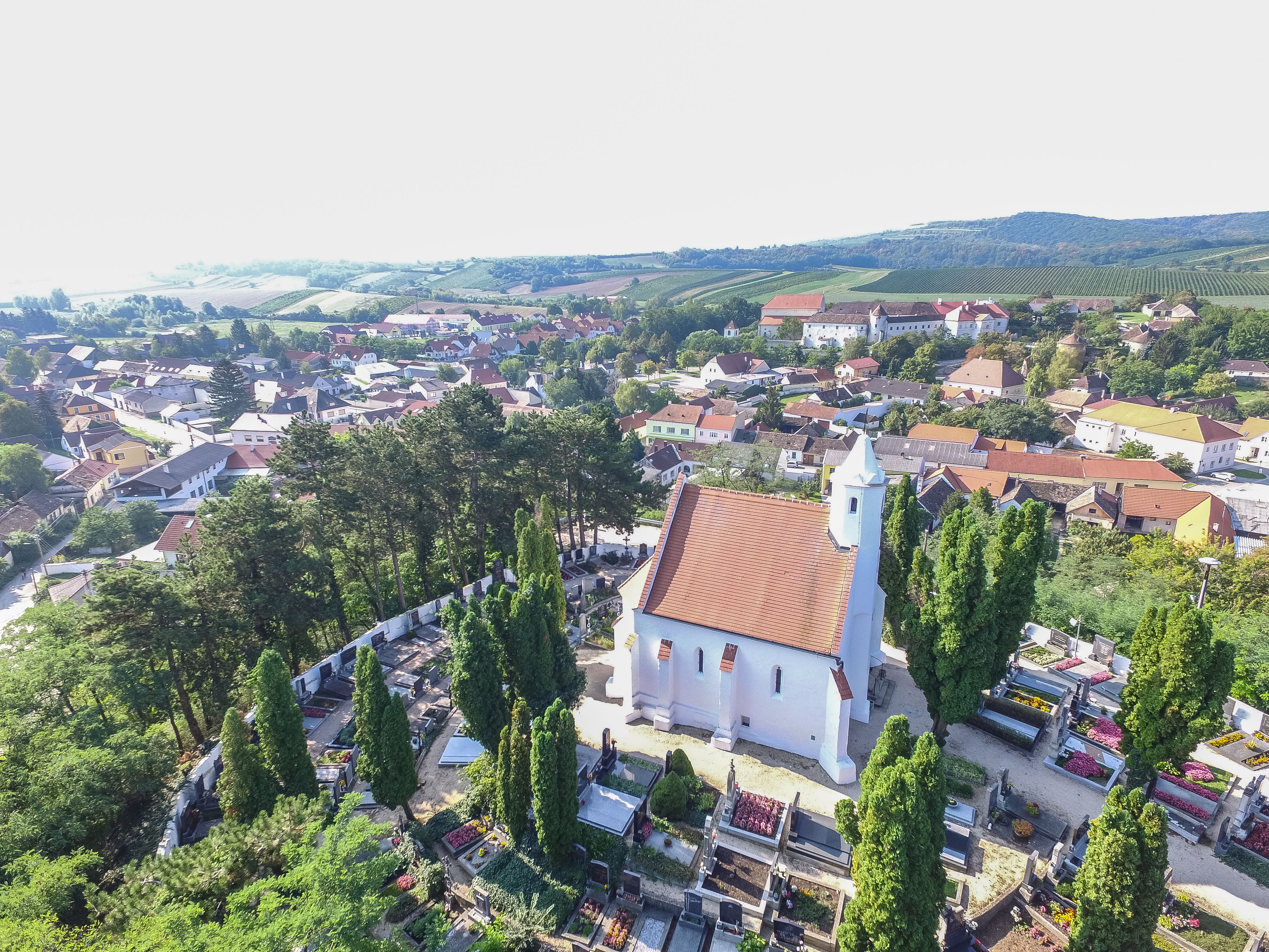 Letecký pohled na kostel s červenou střechou, obklopený hřbitovem a stromy, ve venkovském prostředí s domy a kopci v pozadí.