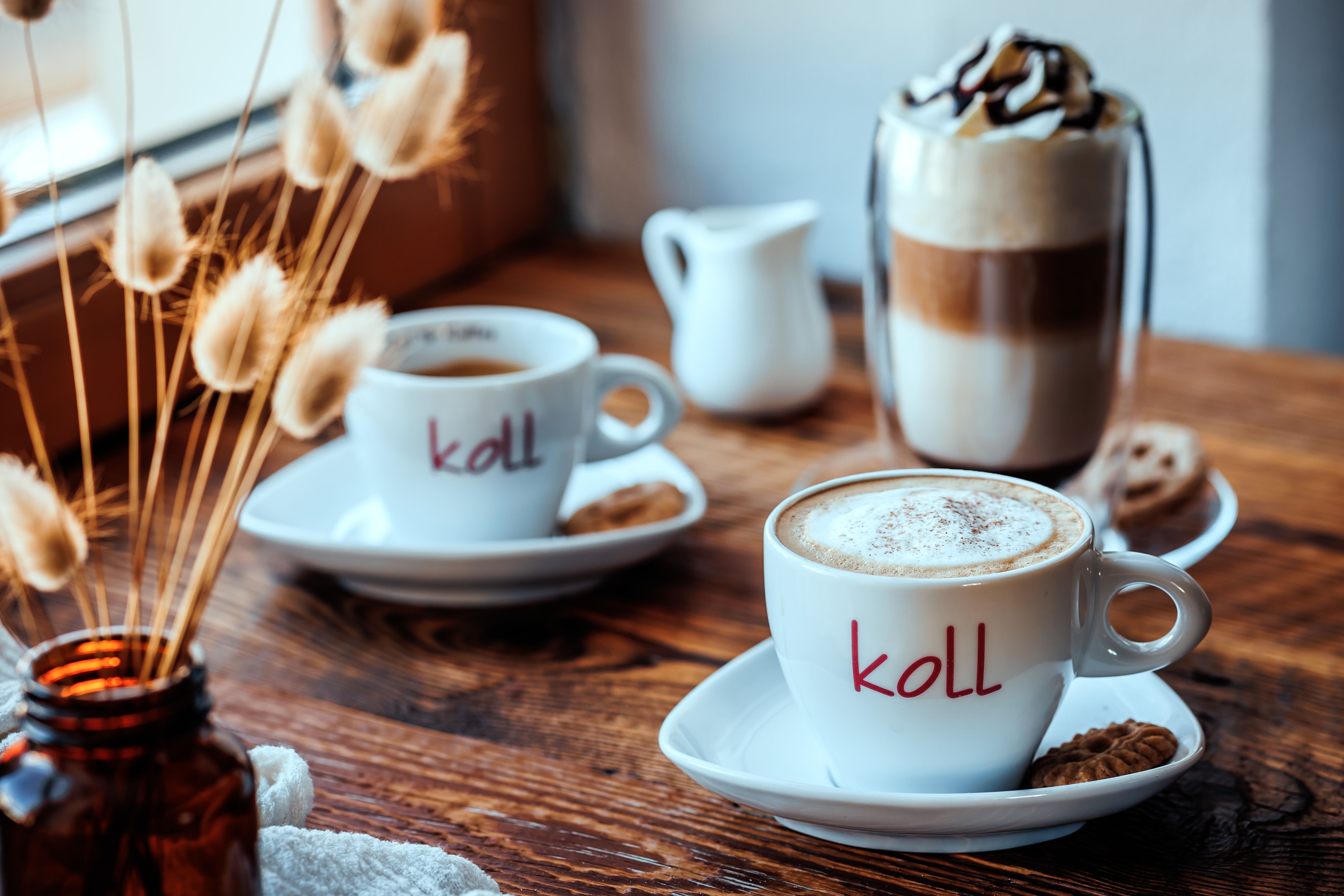 Dva šálky kávy na dřevěném stole, jeden s mléčnou pěnou, sklenice latte macchiato a vedle ní džbánek.