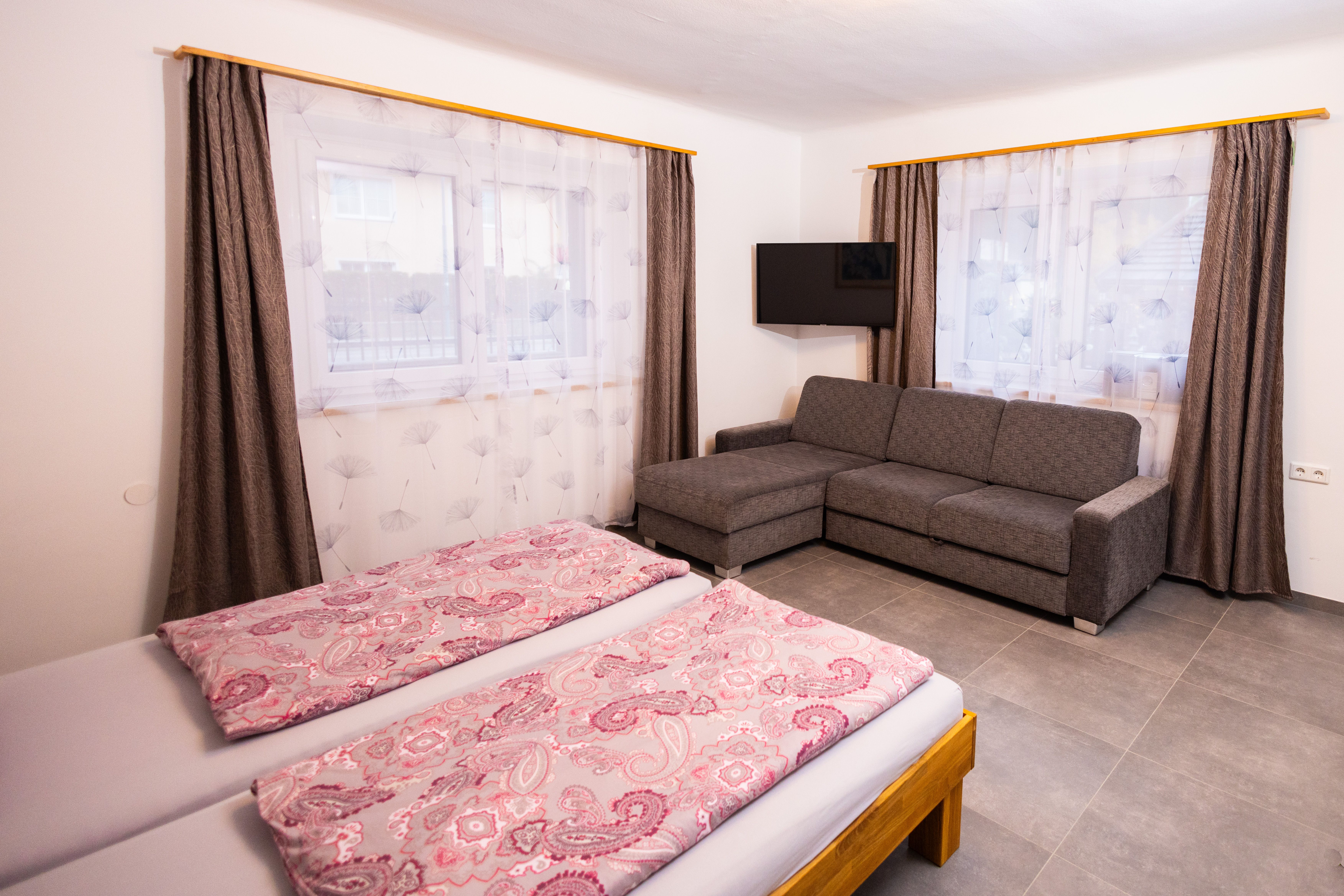 Jedna ložnice s manželskou postelí, šedou pohovkou, TV a dvěma okny se závěsy.