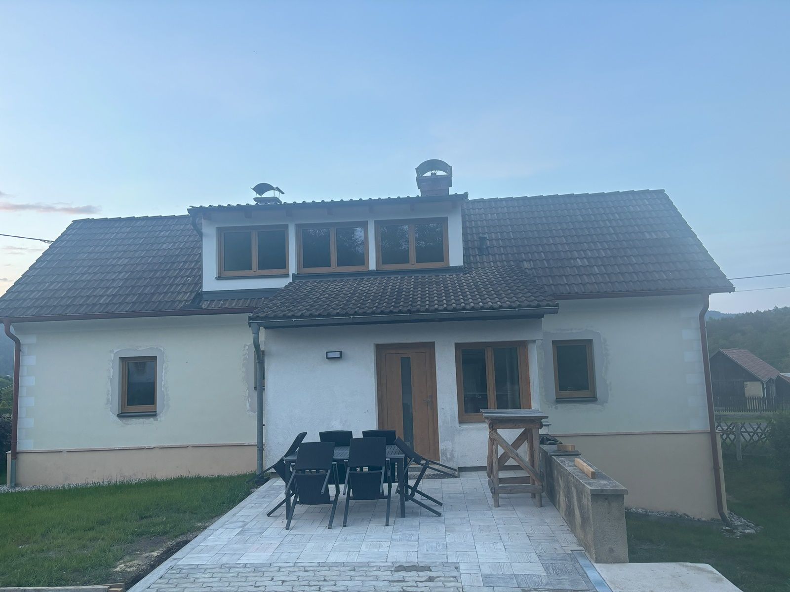 Dvoupodlažní rekreační dům se šindelovou střechou a terasou, obklopený trávníkem a venkovským prostředím.