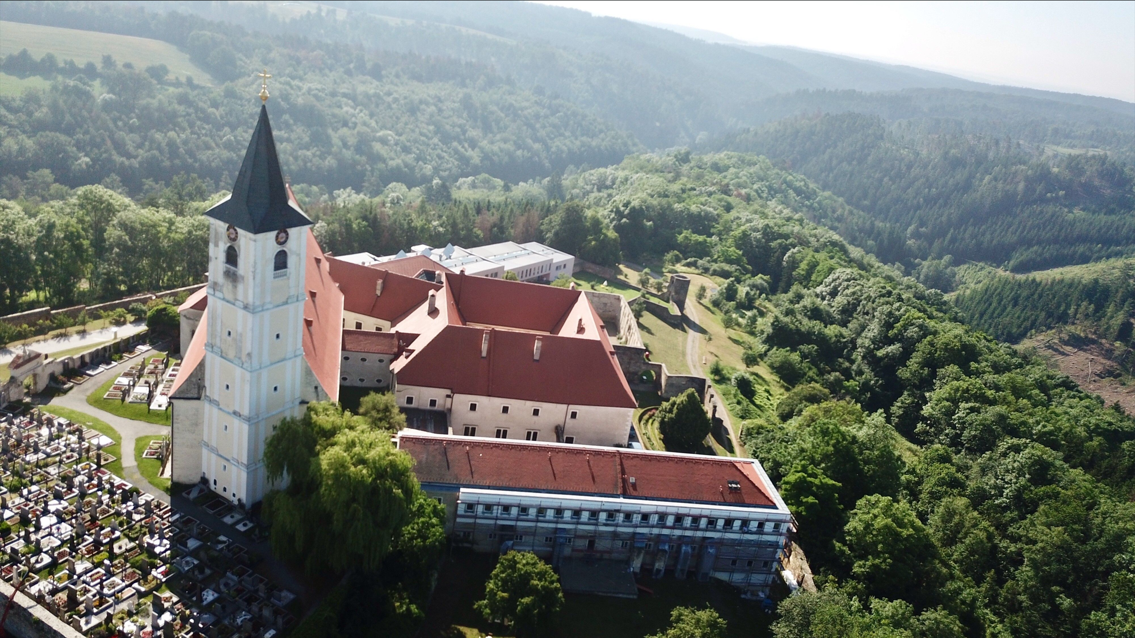 Letecký pohled na klášter s červenou střechou a hřbitovem vedle něj, obklopený zalesněnými kopci.