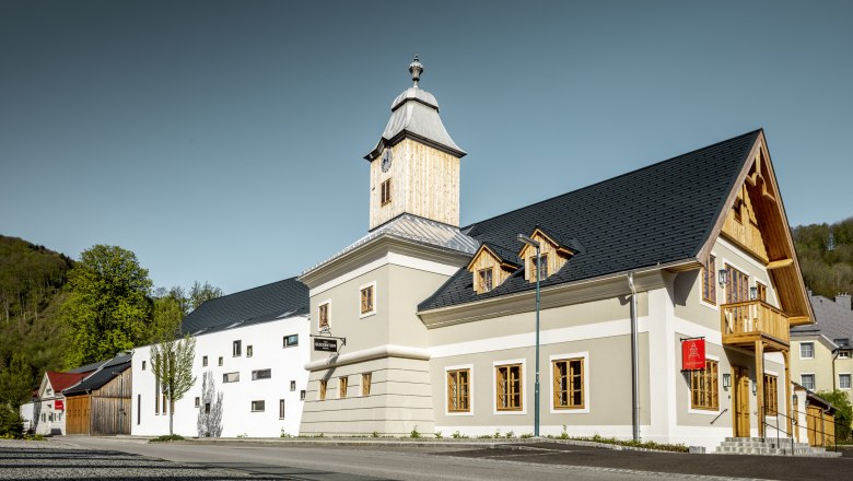 Moderní budova se zvonicí a dřevěným obložením, obklopená stromy a jasnou oblohou.