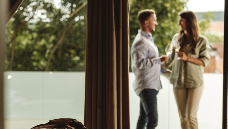 P&aacute;r stoj&iacute; na balkoně a drž&iacute; sklenky v&iacute;na, v popřed&iacute; lež&iacute; na posteli cestovn&iacute; ta&scaron;ka.