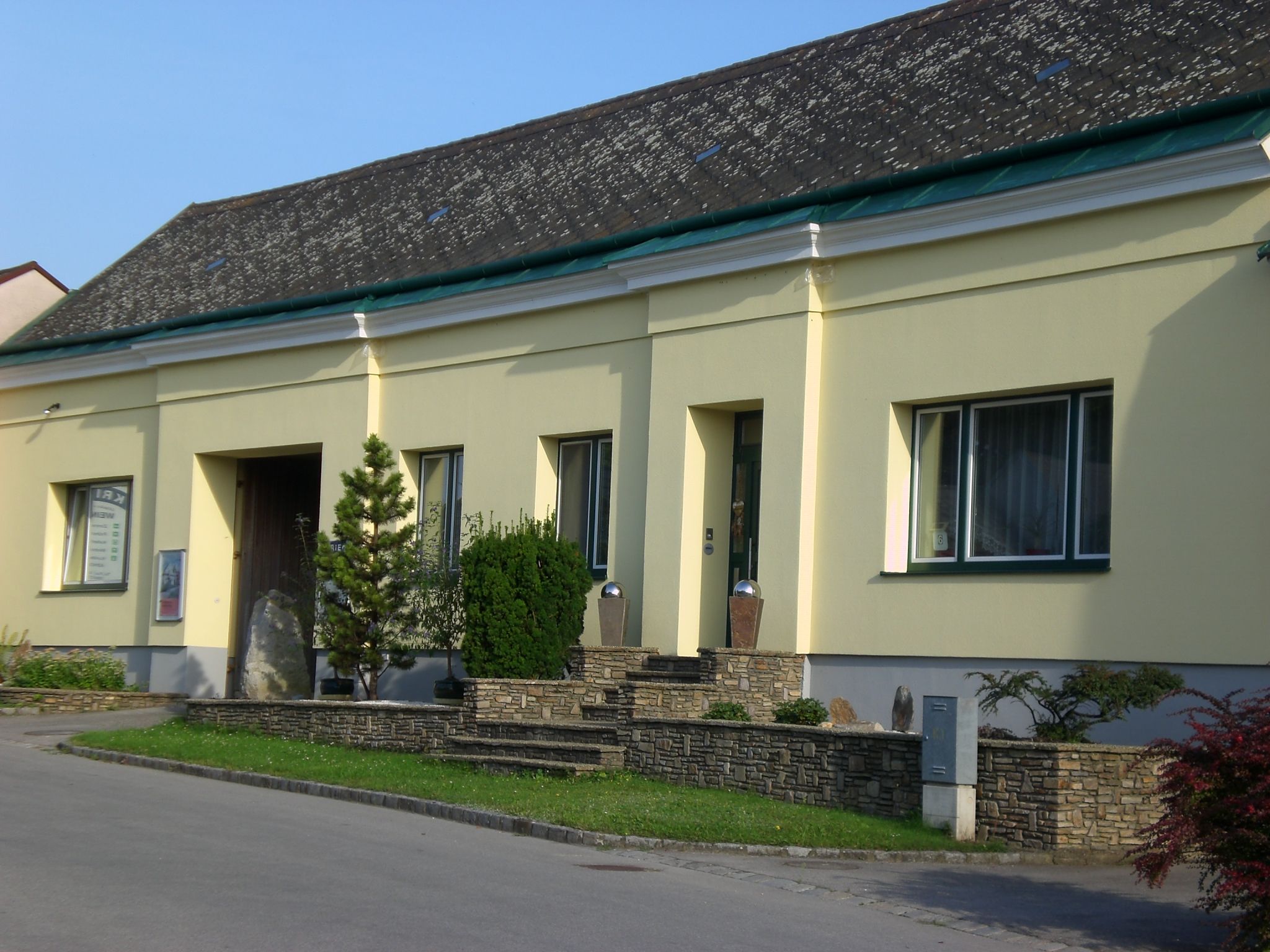 Žlutá budova s kamennou zdí a rostlinami před vchodem.