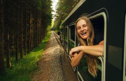 Eine lächelnde Frau lehnt sich aus dem Fenster eines fahrenden Zuges, der durch einen dichten Wald im Waldviertel fährt.